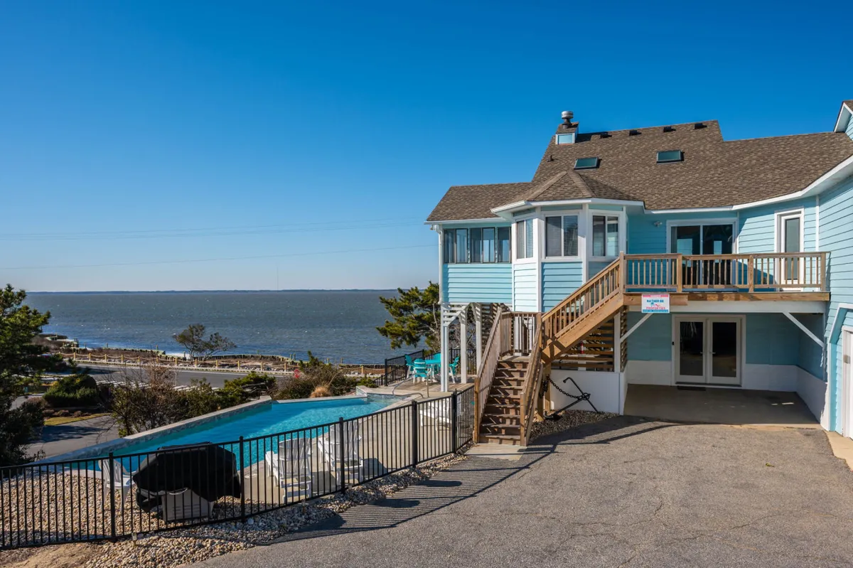 A stunning exterior view featuring an infinity-edge pool in the foreground, the house with large windows and a spacious deck behind it, and expansive waterfront views stretching into the distance. The deck offers space for lounging and socializing, perfect for enjoying sunsets or poolside relaxation.