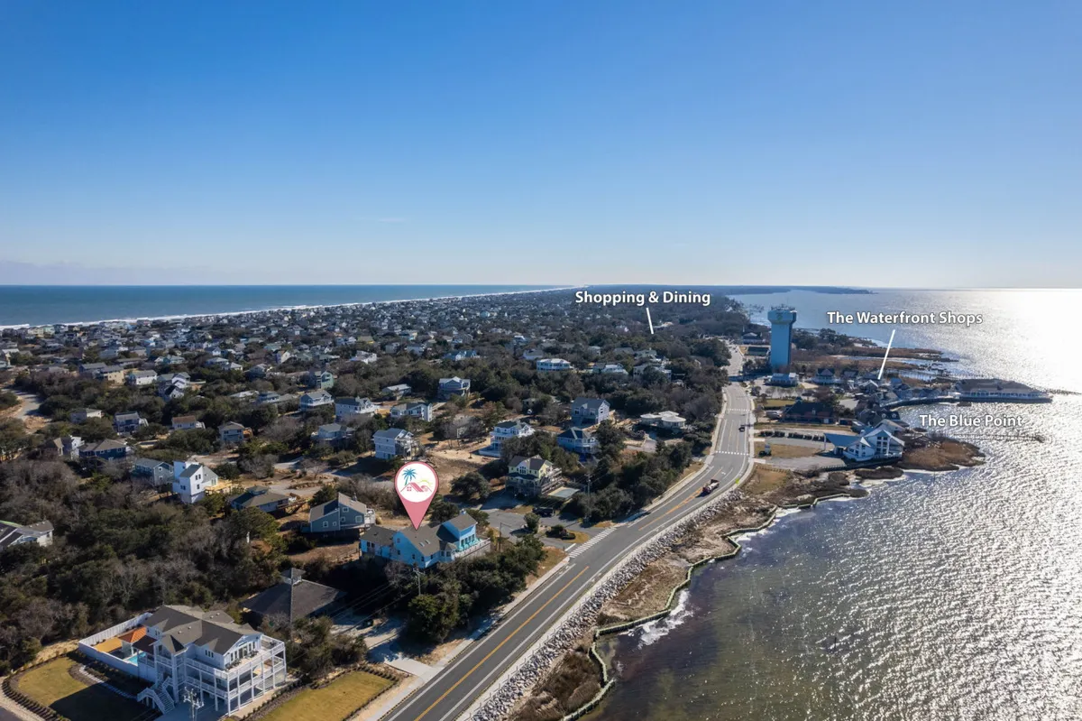 A wide-angle exterior shot of the neighborhood, highlighting the proximity of the waterfront, a cluster of houses, nearby shops, and easy access to the road for exploring local attractions.