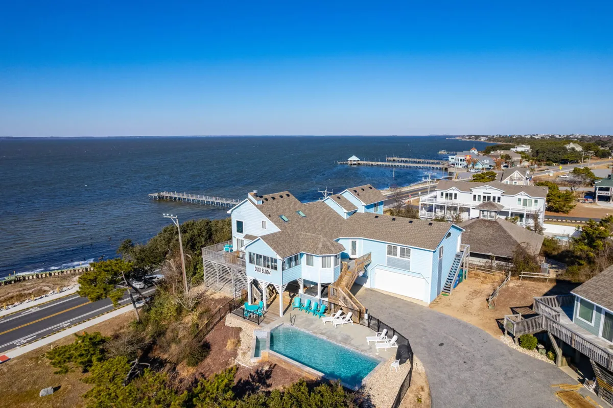 The image showcases a spacious and elegant single-family house perched above the scenic Currituck Sound in Duck, North Carolina. An inviting infinity pool stretches across the foreground, with lounge chairs for sunbathing. Beyond the pool, the water shimmers under the late afternoon light, a private pier extends into the sound, and the expansive deck hints at perfect spots for barbecues and relaxation. The overall mood is tranquil and luxurious, evoking a sense of escape and leisurely waterfront living.