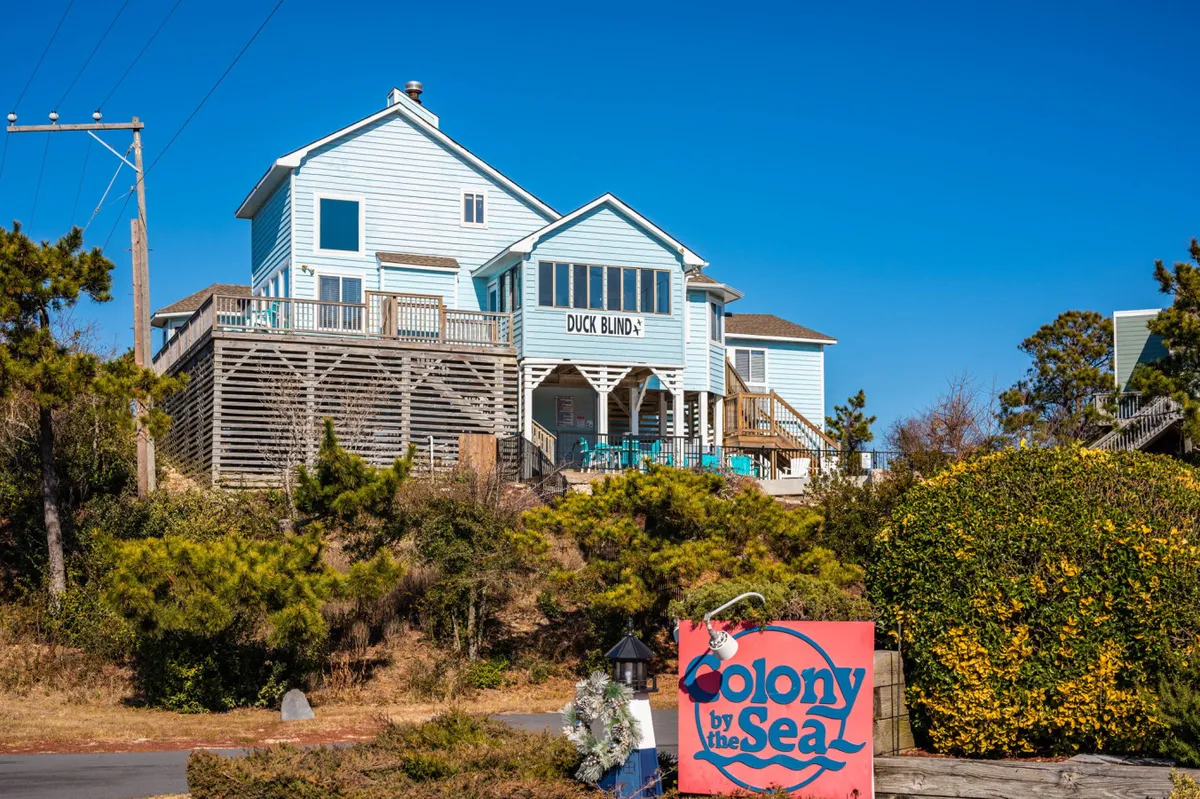 A welcoming shot of Duck Blind's exterior, showing the home, the sign for Colony by the Sea, and lush greenery. The spacious deck and yard are visible, suggesting ample space for pets and their owners to relax and play outdoors.