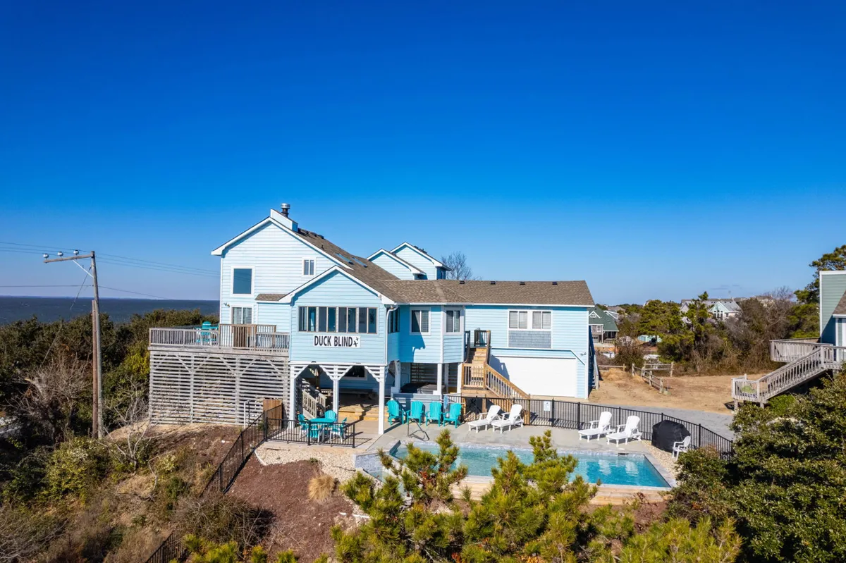A cheerful backyard scene showcases a sparkling swimming pool, inviting outdoor seating, and the welcoming facade of the 'DUCK BLIND' house. The deck area is ideal for barbecues, lounging, and family gatherings, with lush greenery adding privacy.