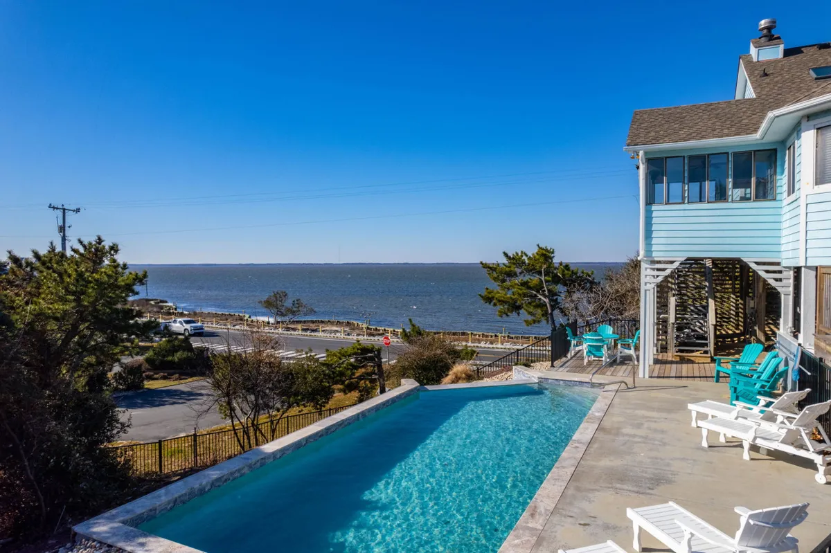 A stunning infinity pool takes center stage, overlooking the ocean with a backdrop of lounge chairs and a contemporary house exterior. The expansive deck area is ideal for sunbathing, outdoor dining, and gatherings, while the pool’s edge blends seamlessly with the water view beyond.