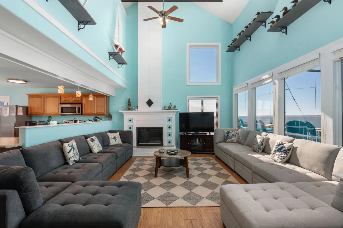 A spacious great room featuring large sectional sofas, a cozy electric fireplace, a coffee table, and expansive floor-to-ceiling windows that fill the space with sunlight and provide panoramic views of the water. Plush seating invites relaxation and the open layout encourages social gatherings.