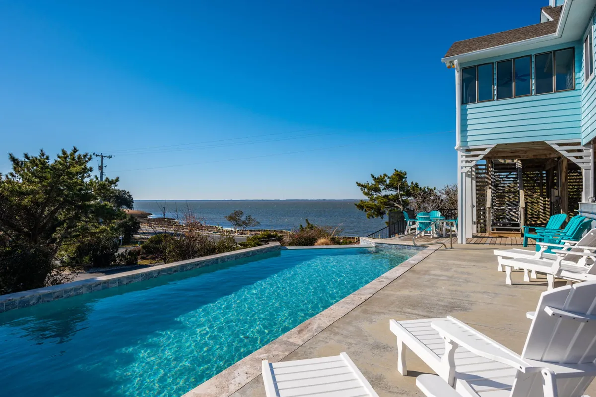 A peaceful exterior view reveals a swimming pool lined with deck chairs, all set against a beautiful waterfront. The tranquil mood is perfect for sunbathing, relaxing with friends, or enjoying a quiet morning coffee by the pool.