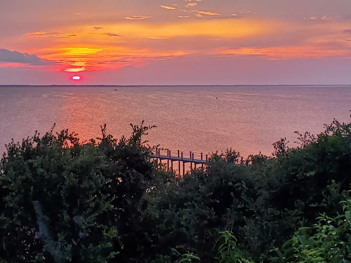 A tranquil sunset scene with a pier stretching over calm water, silhouetted vegetation, and the sun dipping below the horizon. The golden hour creates a magical, serene atmosphere ideal for photography or quiet reflection by the water.