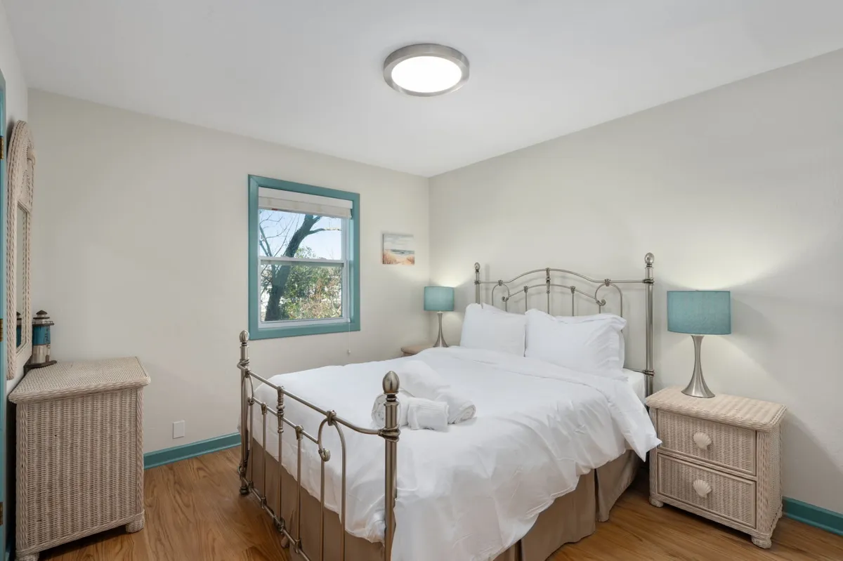 This image features a serene bedroom with a bed dressed in crisp white linens, blue bedside lamps, and a woven dresser. Natural light floods the space, creating a calming atmosphere perfect for relaxation or reading. The overall feel is tranquil and inviting, ideal for restful nights and slow mornings.